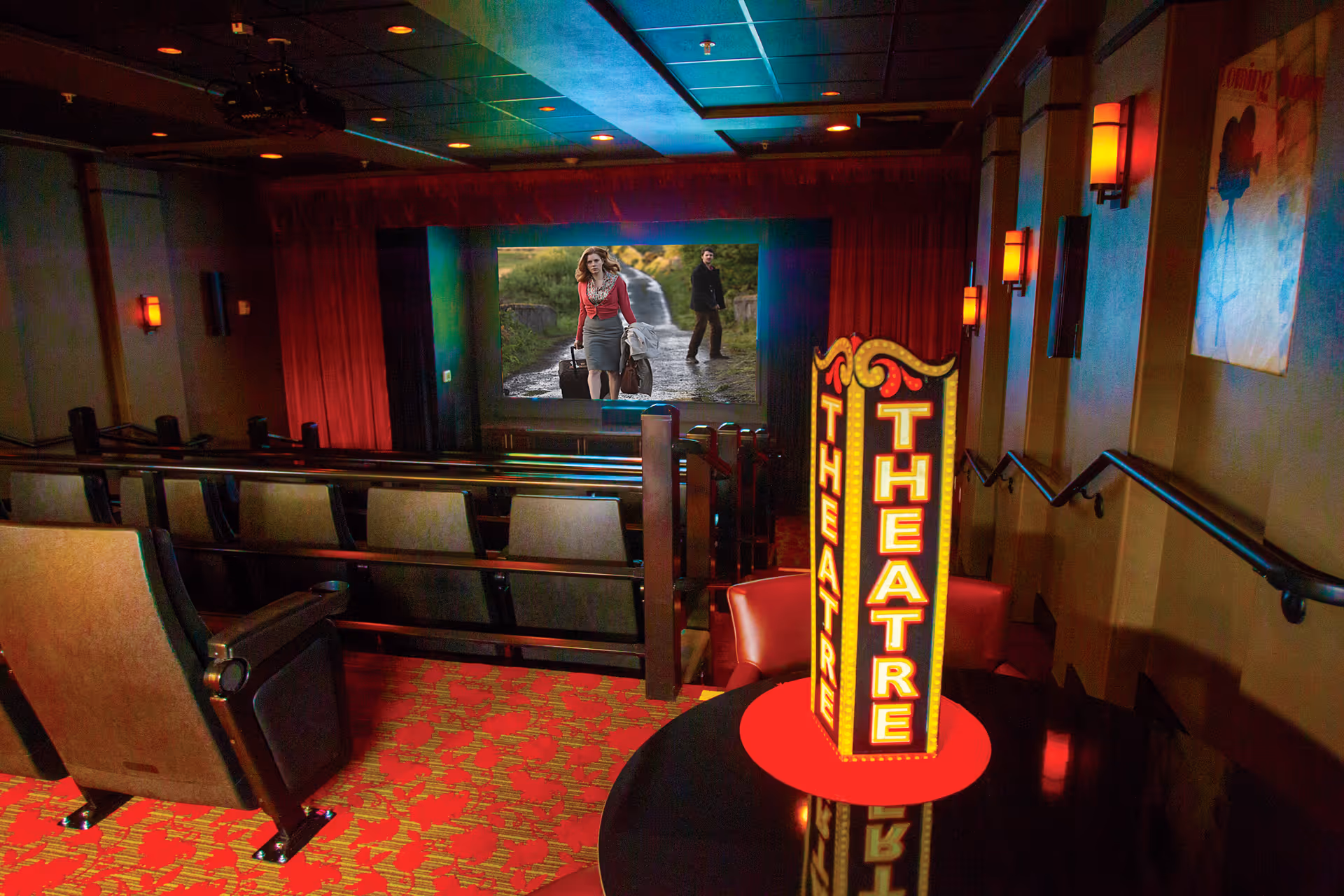 Interior view of a small theater room with rows of seats facing a large screen showing a movie scene with a woman and a man. The room has red patterned carpet, wall sconces with warm lighting, and a decorative illuminated sign that says 'THEATRE' on a round table near the entrance.
