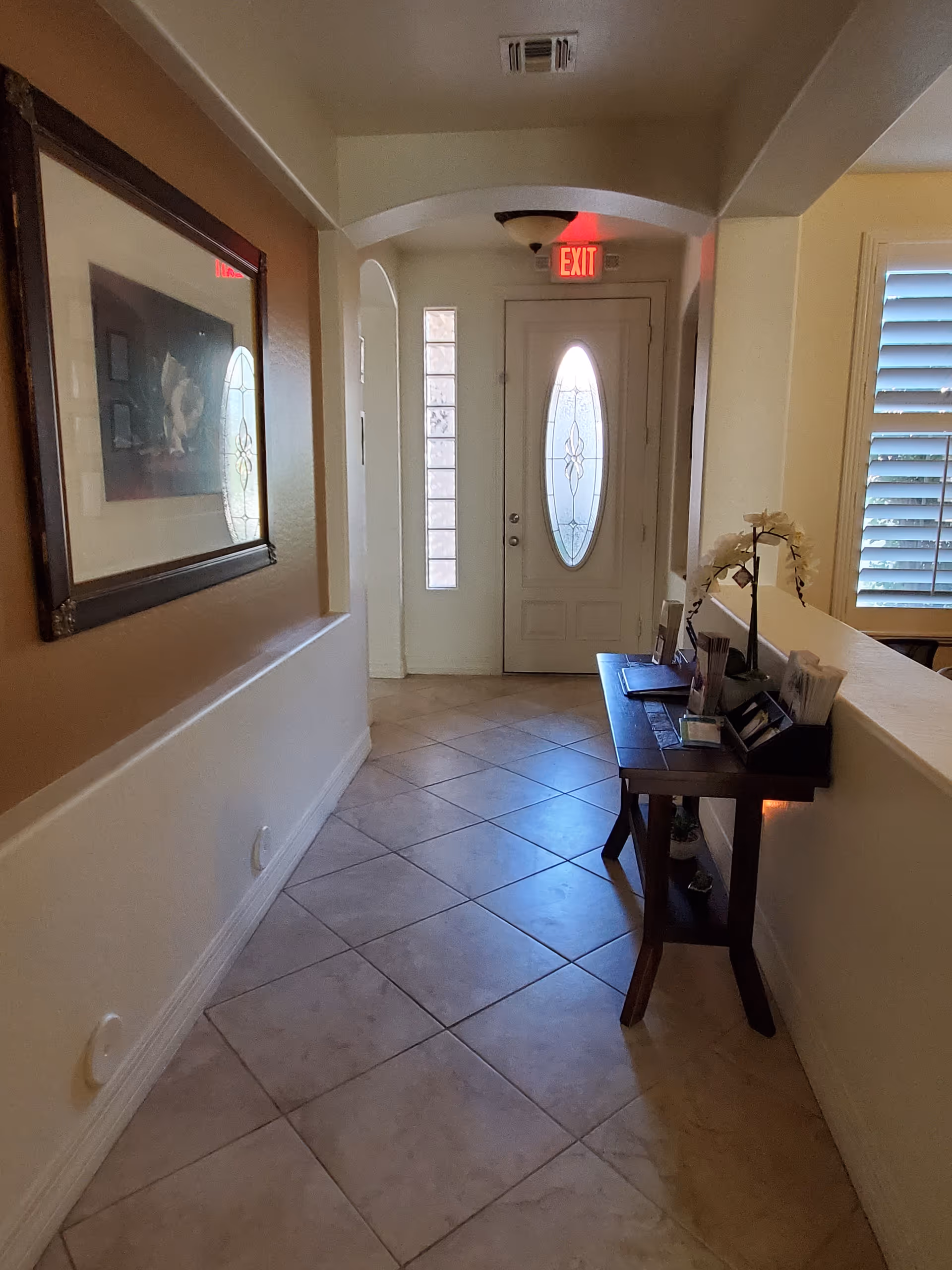 A hallway with beige tiled floor leading to a white front door with an oval glass window. There is an exit sign above the door. On the right side, there is a wooden table with decorative items including a white orchid plant and some brochures or pamphlets. On the left wall, there is a framed picture. The hallway is lit by natural light coming through the window blinds on the right.