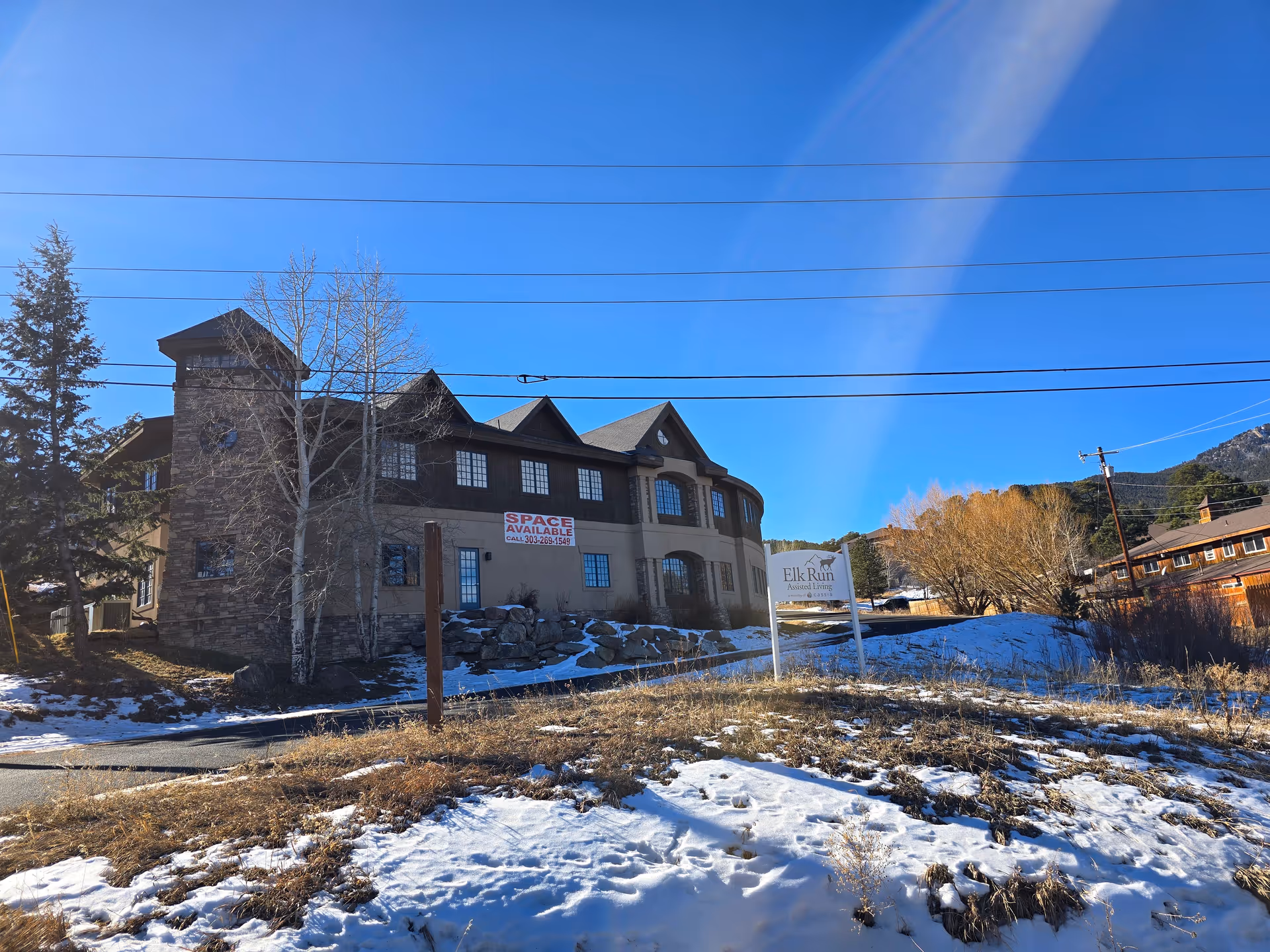 Exterior view of Elk Run Assisted Living facility on a clear day with blue sky. The building has a stone and beige facade with multiple windows and a sign in front that reads 'Elk Run Assisted Living'. There is some snow on the ground and dry grass in the foreground, with trees and mountains in the background.