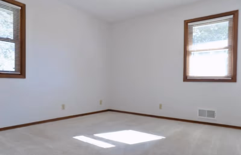 Empty room with white walls, beige carpet, two windows with wooden blinds, and wooden trim around the windows and baseboards.