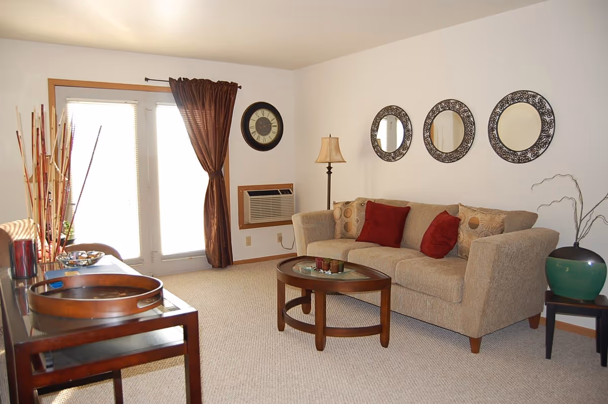 A cozy living room with a beige sofa adorned with red and patterned cushions, a round wooden coffee table with decorative items, a floor lamp, and three decorative round mirrors on the wall. There is a wooden side table with a large green vase and a small table with a wooden tray and other decorative items. French doors with brown curtains allow natural light into the room.