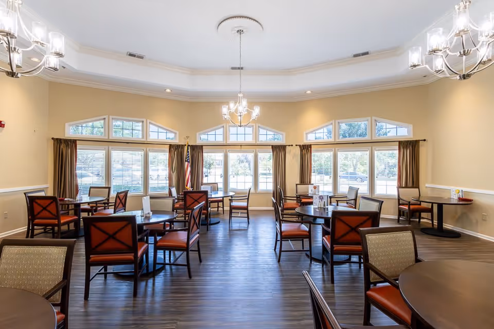 A bright dining room with multiple round tables and chairs arranged neatly. Large windows with brown curtains allow natural light to fill the room. The ceiling features elegant chandeliers and recessed lighting. The floor is dark wood, and an American flag is visible near the windows.