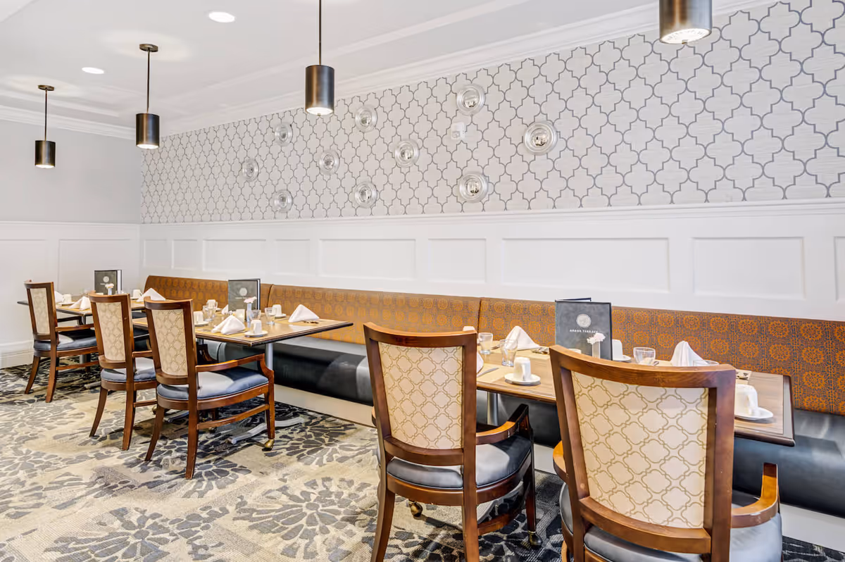 A dining room in Tappan assisted living nursing home featuring wooden tables set with white napkins, cups, and menus. The room has patterned wallpaper, a long cushioned bench along the wall, wooden chairs with upholstered backs and seats, pendant lights hanging from the ceiling, and a patterned carpet.