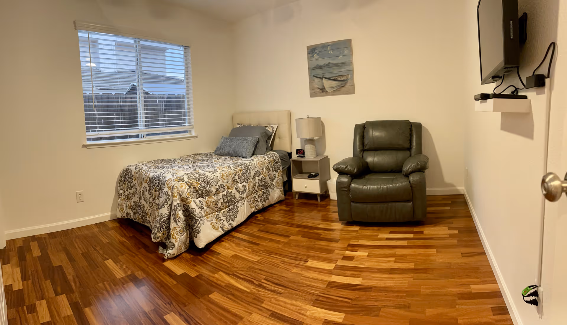 A small bedroom with a single bed and patterned bedspread, a nightstand with a lamp, a green recliner, a wall-mounted TV, and hardwood floors.