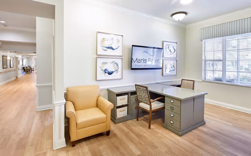 A bright office area in a senior living facility with a large window letting in natural light. The room features a gray desk with two chairs, a beige armchair, and abstract artwork on the wall. A flat screen TV mounted on the wall displays the text 'Maris Pointe Eagle Senior Living.' The floor is light wood, and the walls are painted a soft cream color.