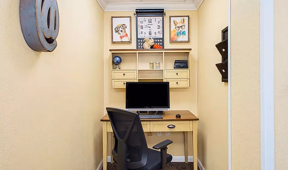 A small narrow office nook with a desk, computer monitor, mesh office chair, wall-mounted shelving, framed animal prints and a clock.