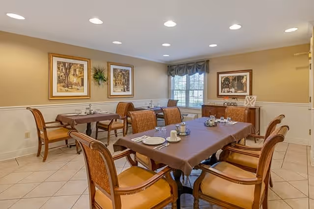 A dining room with two tables covered in brown tablecloths, each set with plates, cups, and utensils. The room has beige walls with white wainscoting, framed artwork, a window with a valance, and wooden chairs with cushioned seats around the tables.