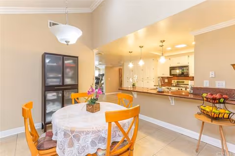 A dining area with a round table covered with a white lace tablecloth and four wooden chairs with orange cushions. A small flower arrangement is on the table. Behind the table is a dark cabinet with frosted glass doors. To the right is a kitchen counter with three hanging pendant lights above it. The kitchen has white cabinets, a microwave, and a stove. A small wooden table with a tiered fruit basket is also visible.