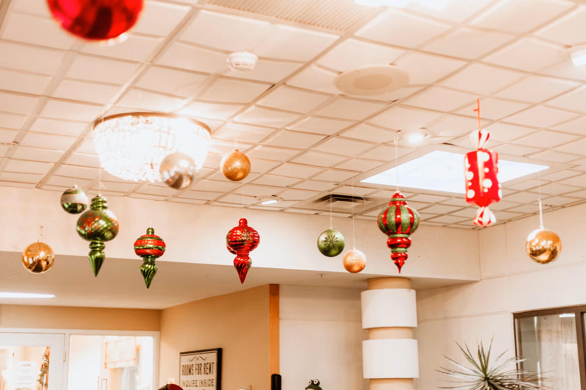 Ceiling decorated with hanging Christmas ornaments in red, green, and gold colors inside a senior living community room with a chandelier and a skylight.