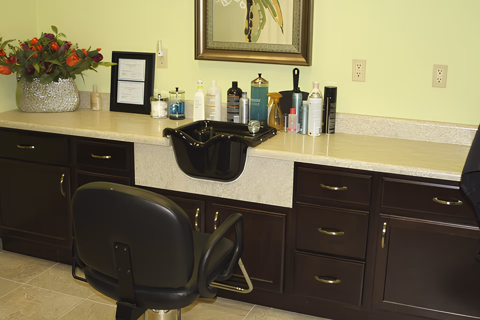 Interior salon station with a black shampoo sink, salon chair, dark wood cabinets and various hair products on the counter.