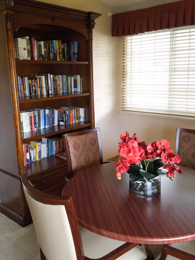 A cozy interior room featuring a round wooden table with a vase of vibrant red flowers in the center, surrounded by upholstered chairs. Behind the table is a tall wooden bookshelf filled with books. A window with white blinds and a red valance lets in natural light.