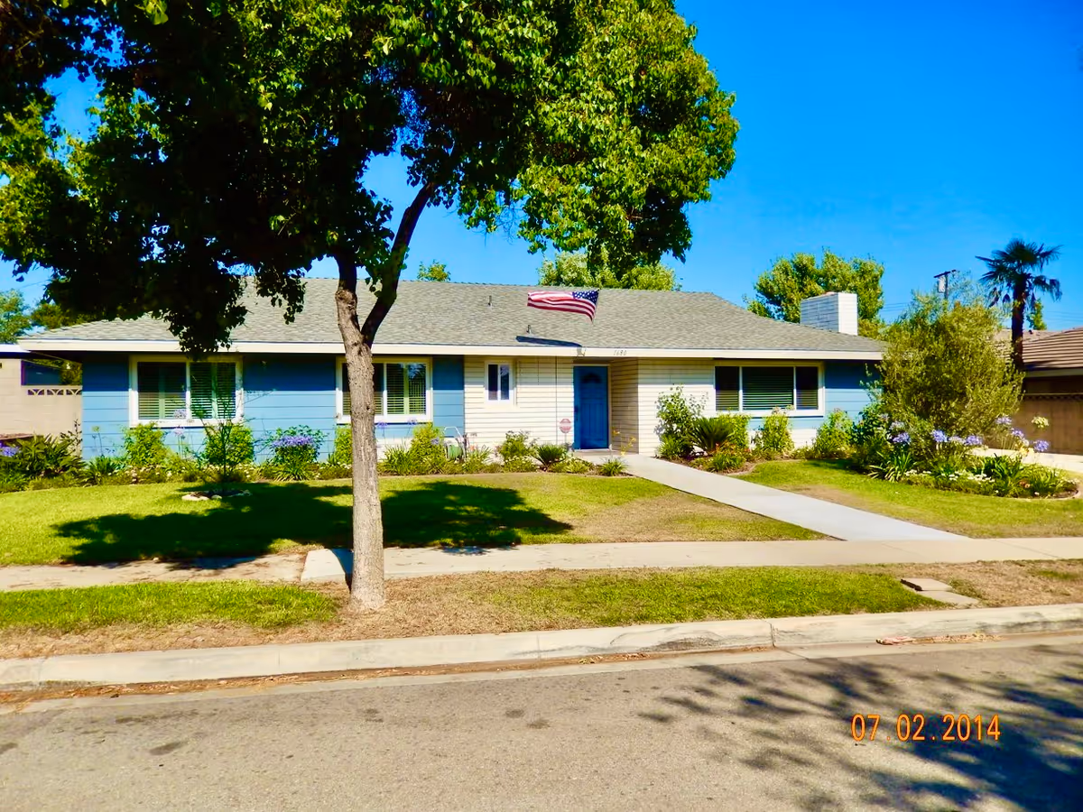 Single-story residential house with blue and beige exterior walls, a gray shingled roof, and an American flag mounted above the front door. The house is surrounded by a well-maintained lawn, trees, and shrubs under a clear blue sky.