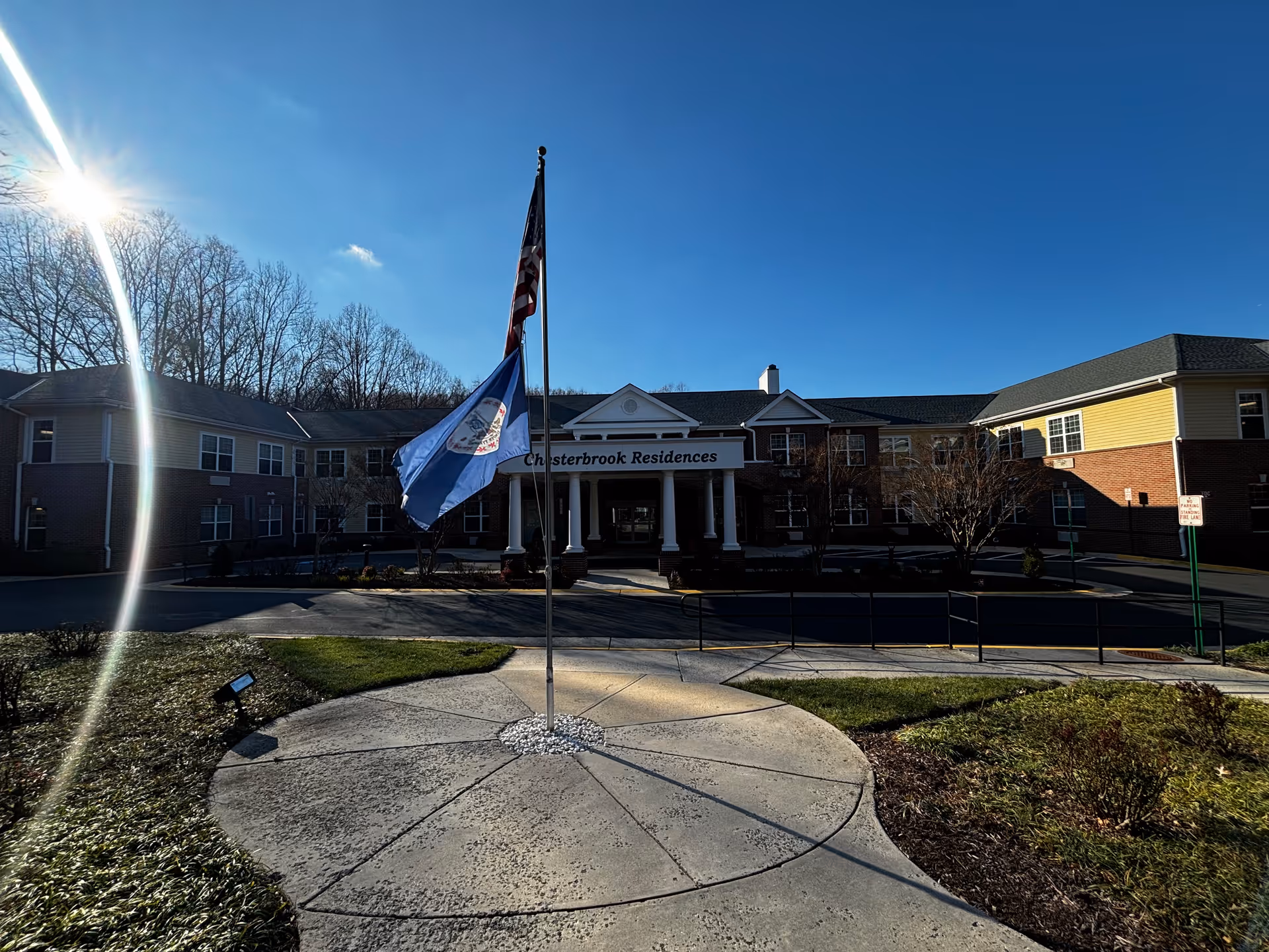 Exterior front view of Chesterbrook Residences building with a circular concrete pathway and two flagpoles displaying the American flag and another flag. The building is two stories with a brick and siding facade, under a clear blue sky with the sun shining brightly.