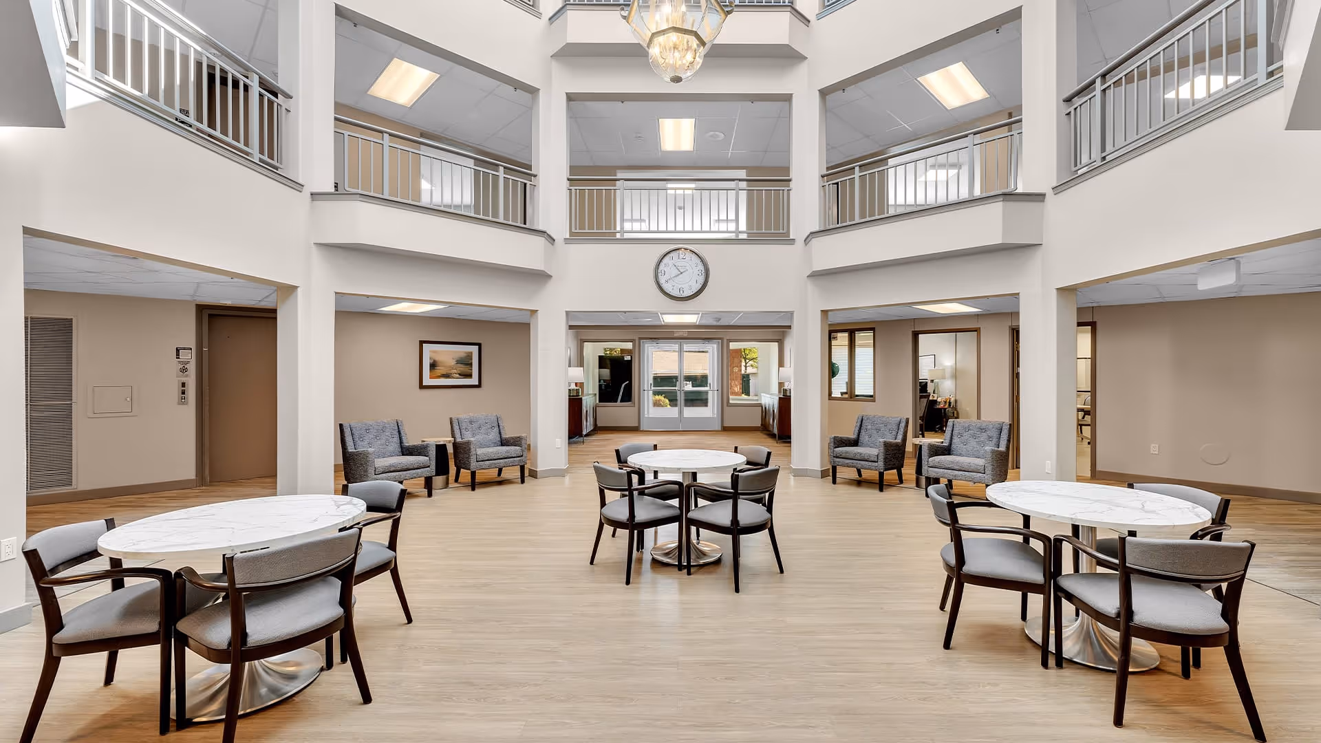 Spacious and well-lit common area in a senior living facility with three round tables each surrounded by four chairs. There are several armchairs arranged along the walls. The room features a high ceiling with a large chandelier and a clock mounted above the entrance doors. The upper level has railings overlooking the area below.