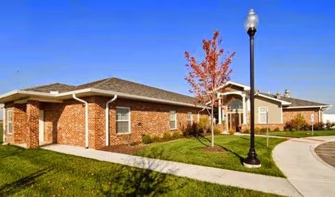 Exterior view of a single-story brick building with a well-maintained lawn, a young tree with red leaves, a sidewalk, and a streetlamp under a clear blue sky.