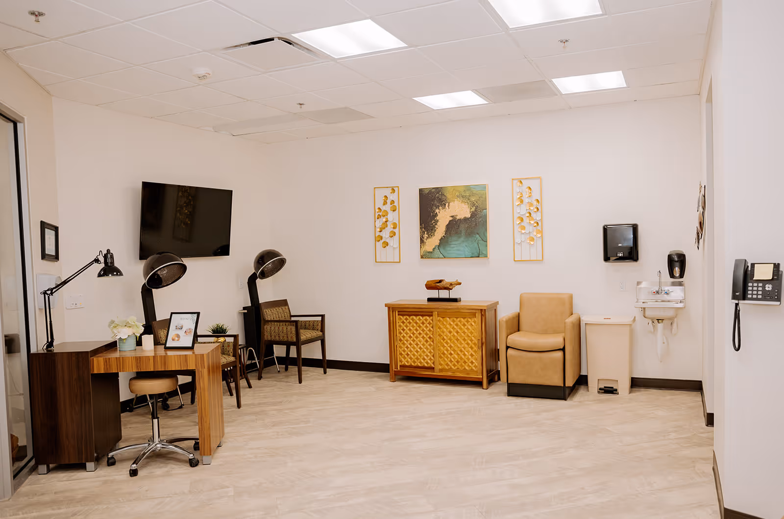 A clean and well-lit interior room with light-colored walls and flooring. The room features a wooden desk with a lamp, a rolling chair, two vintage hair dryer chairs, a wall-mounted flat screen TV, a small wooden cabinet with decorative items, a tan armchair, a wall-mounted sink with soap dispenser and paper towel holder, a trash bin, and a wall phone. Three pieces of framed artwork hang on the wall above the cabinet and armchair.