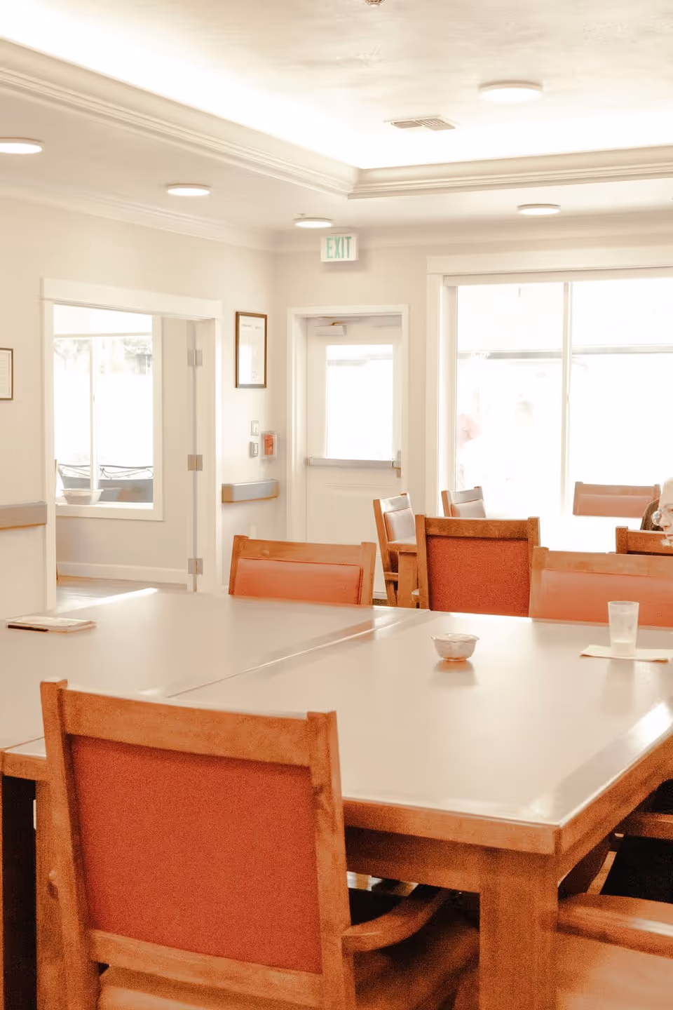 A bright dining room with wooden tables and chairs featuring red cushions. There is a large window letting in natural light, an exit door, and a hallway leading to another room. The ceiling has recessed lighting and crown molding.