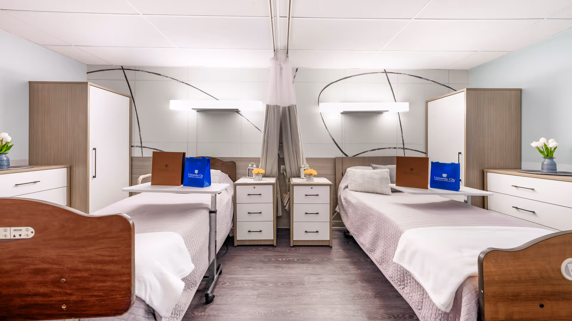 A bright and clean shared bedroom in a healthcare facility with two hospital-style beds separated by a curtain. Each bed has a bedside table with drawers, a small flower vase, and an overbed table holding a brown folder and a blue bag labeled 'University City'. The room features modern wood furniture, light-colored walls with abstract designs, and a wood floor.