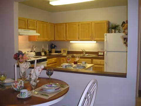 View of a kitchen with wooden cabinets, a white refrigerator, a stove with a white hood, and a countertop with a sink. In the foreground, there is a dining table set with plates, cups, glasses, and napkins, along with a floral centerpiece.