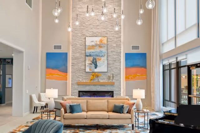 A spacious and well-lit living room area with a beige sofa adorned with blue and brown cushions, two white table lamps on side tables, a modern fireplace with a stone wall backdrop, abstract paintings on either side, and large windows letting in natural light. A black grand piano is visible on the right side.