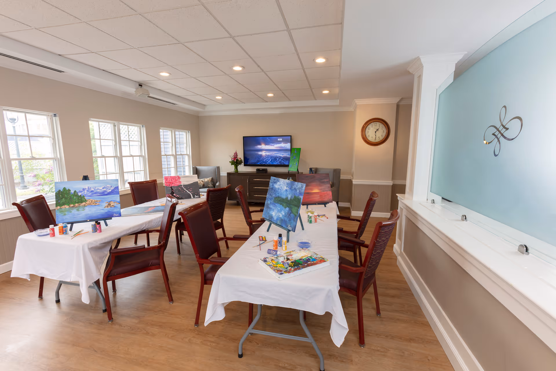 A bright, spacious activity room with large windows letting in natural light. There are two tables covered with white tablecloths, each set up with painting supplies including canvases on easels, paint bottles, brushes, and palettes. Several chairs are arranged around the tables. In the background, a flat-screen TV is mounted on a cabinet with a flower vase beside it. A clock is mounted on the wall, and a decorative frosted glass panel with a design is on the right side.