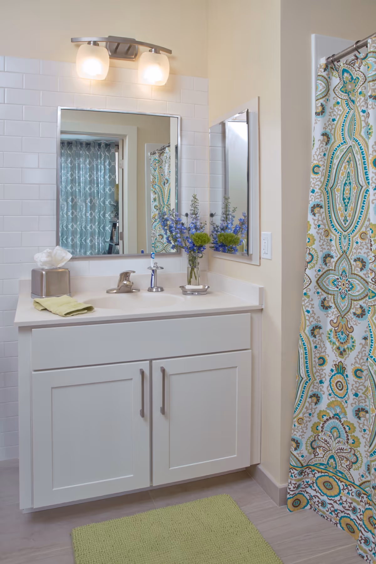 A clean and bright bathroom vanity area with a white cabinet, a sink with a silver faucet, a rectangular mirror above the sink, and a light fixture with two frosted glass shades. There is a tissue box, a folded green towel, and a small vase with purple and green flowers on the countertop. To the right, a colorful patterned shower curtain with blue, green, and beige designs is visible. A green bath mat is on the floor in front of the vanity.