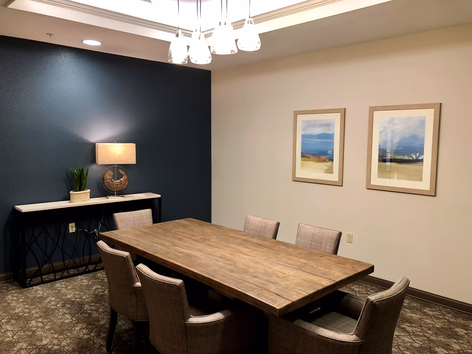 A small meeting or dining room with a wooden rectangular table surrounded by six upholstered chairs. The room has a dark blue accent wall with a console table holding a lamp and a plant. Two framed landscape paintings hang on the adjacent light-colored wall. A modern ceiling light fixture with multiple bulbs hangs above the table.