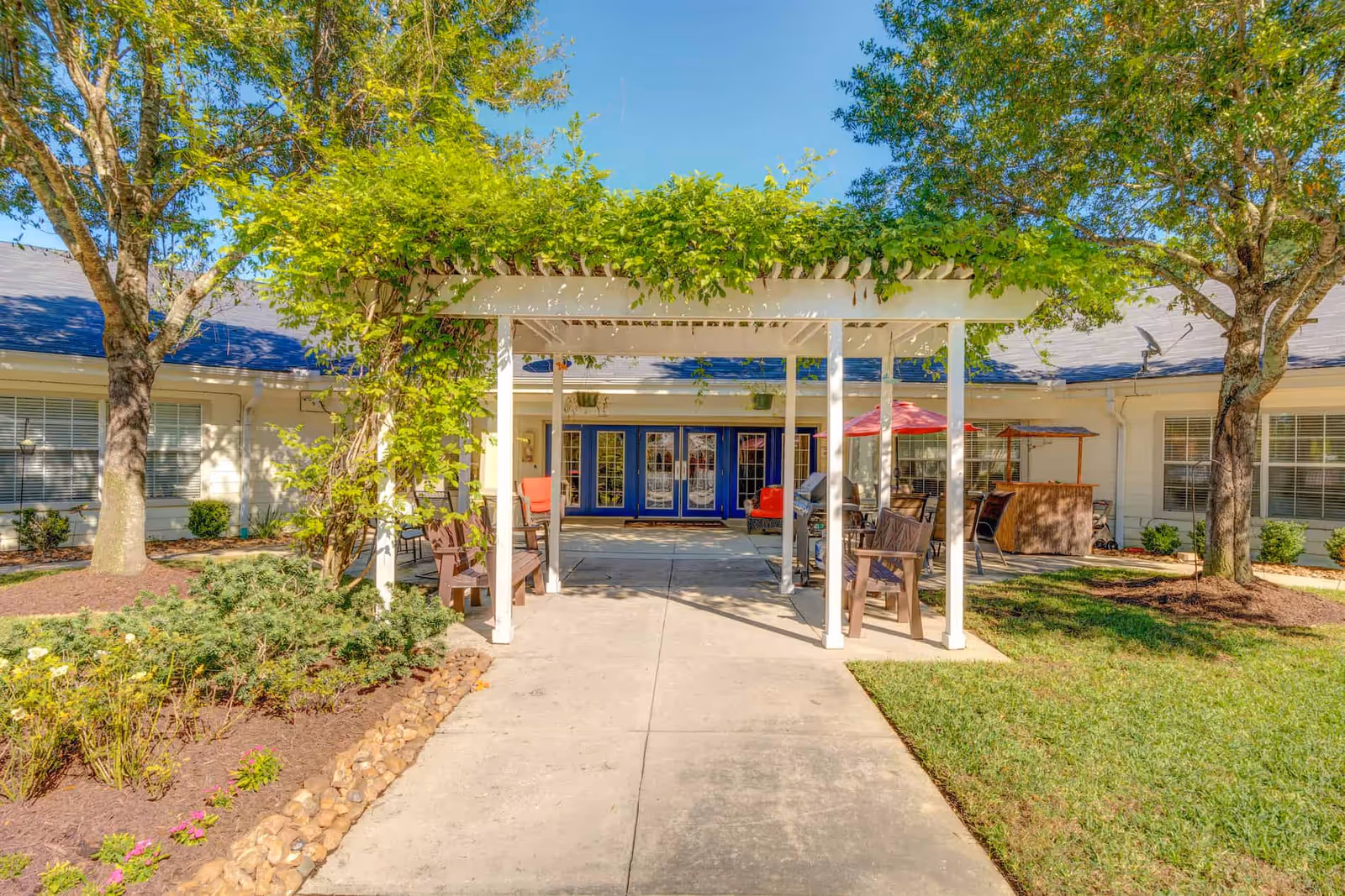 Courtyard entrance with a white pergola, benches, patio seating and greenery leading to the building's blue double doors.