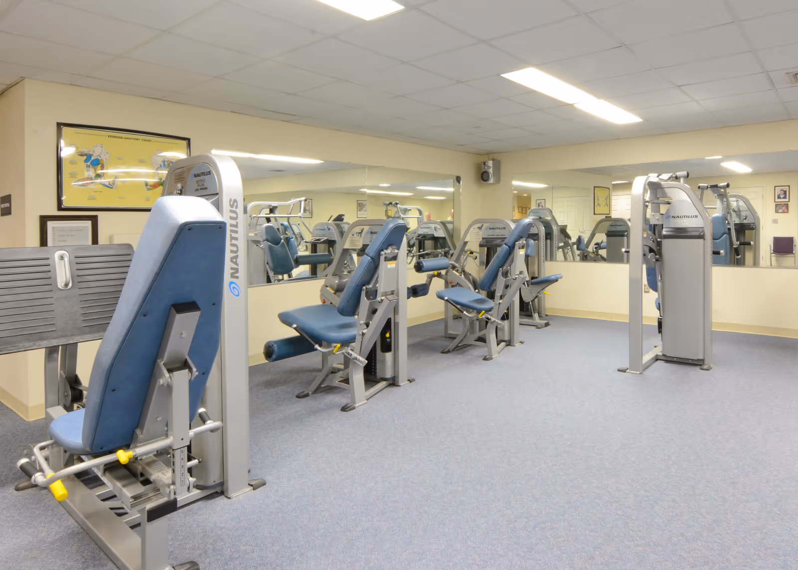 A fitness room with several Nautilus exercise machines featuring blue padded seats and backrests. The room has a carpeted floor, beige walls, and a large mirror covering one wall. There are framed posters and certificates on the walls.