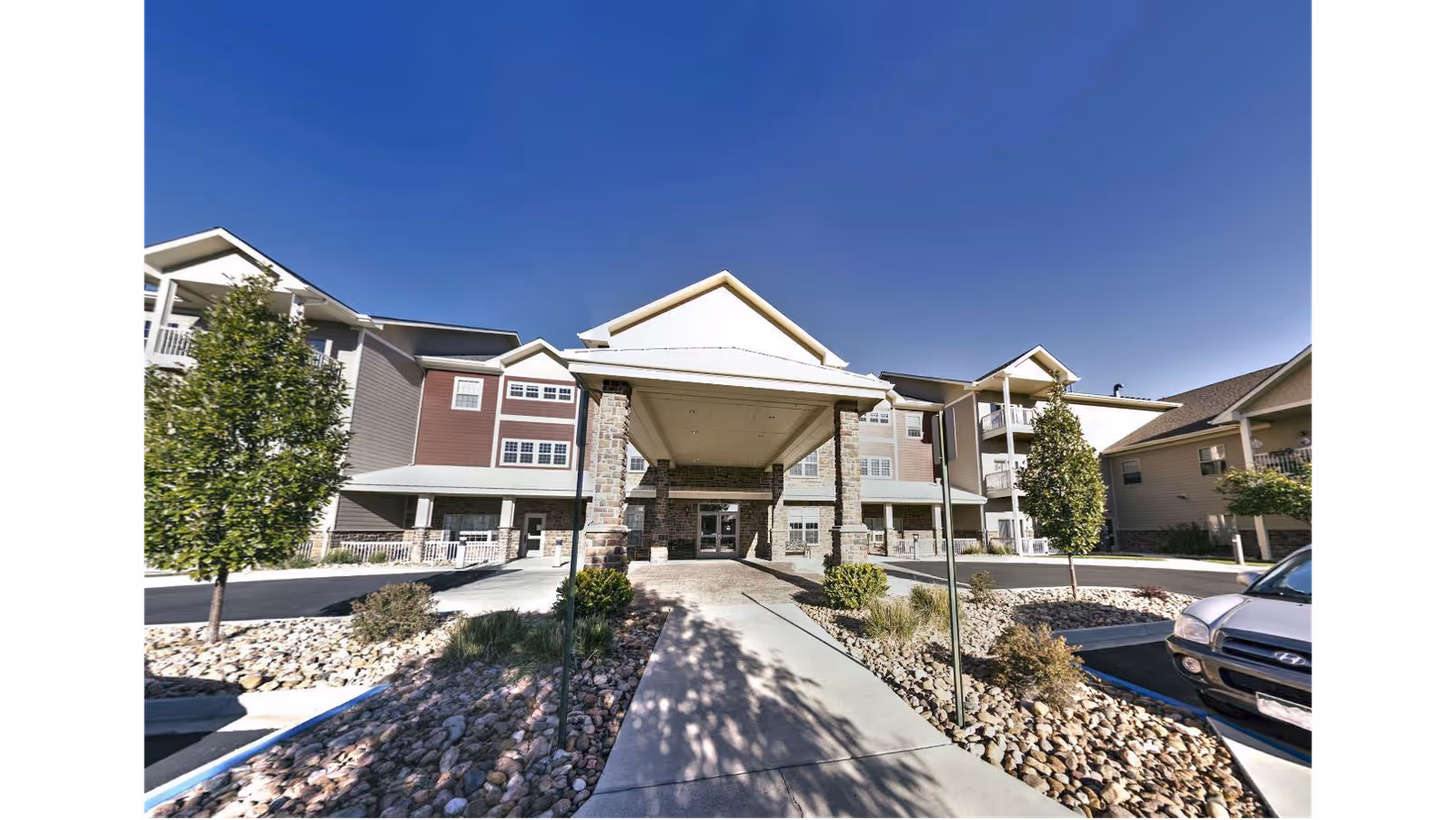 Front exterior view of Primrose Retirement Community of Pueblo showing a covered entrance with stone pillars, landscaped areas with rocks and small trees, and a clear blue sky.