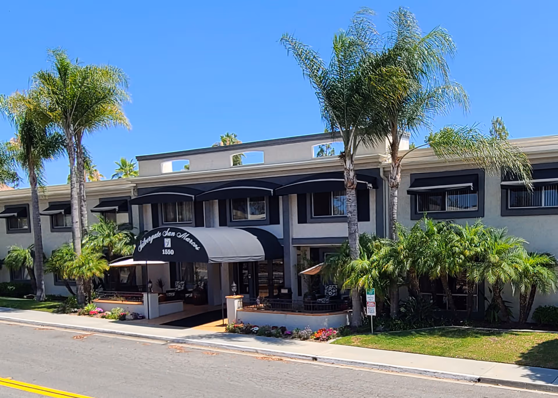 Exterior front view of Silvergate San Marcos facility with palm trees and a black awning over the entrance displaying the facility name and address number 1550, under a clear blue sky.