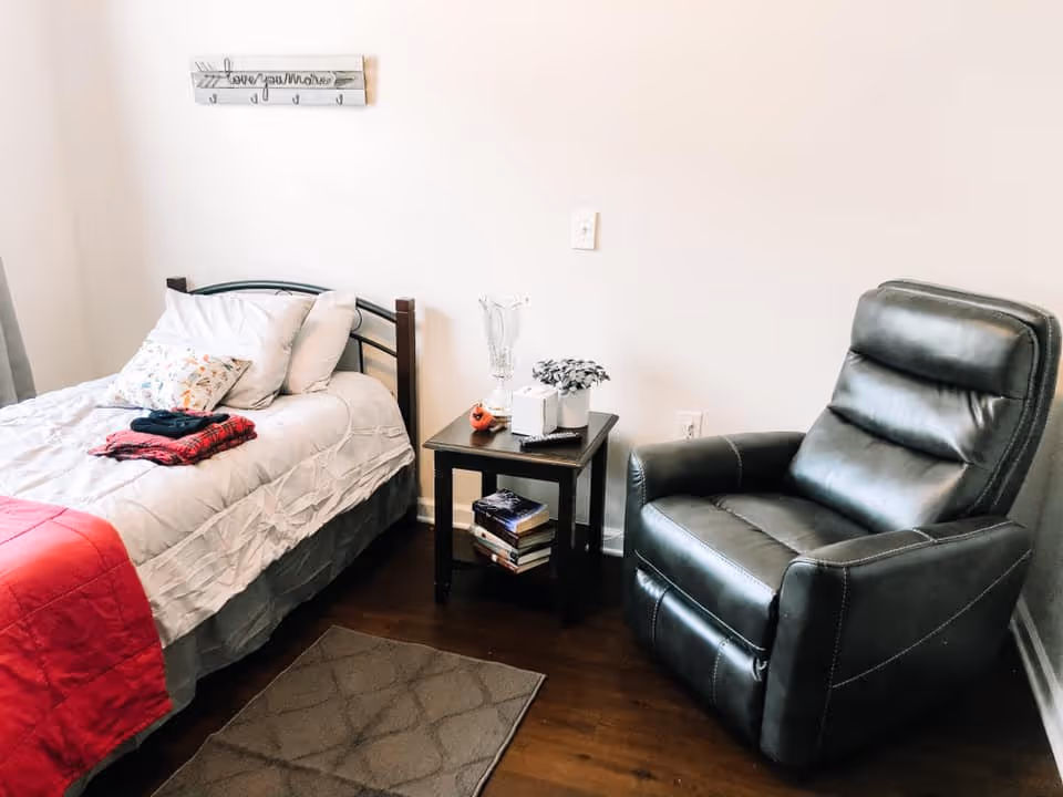 A cozy bedroom with a single bed made with white bedding and a red blanket at the foot. There are pillows on the bed and folded clothes placed on top. Next to the bed is a small dark wooden side table holding a glass vase, a small plant, a remote control, and some books on the lower shelf. To the right of the table is a black leather recliner chair. The floor is wooden with a small gray rug in front of the bed. A wall decoration above the bed reads 'love you more'.