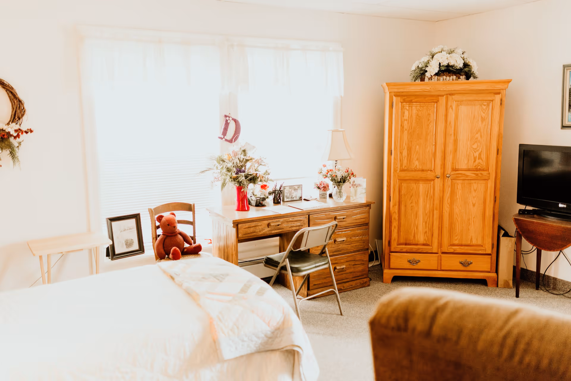 A cozy bedroom with a bed covered in a white quilt, a wooden desk with flowers and a lamp, a chair, a wooden wardrobe, a TV on a small table, and a teddy bear sitting on a chair near the window with sheer white curtains.