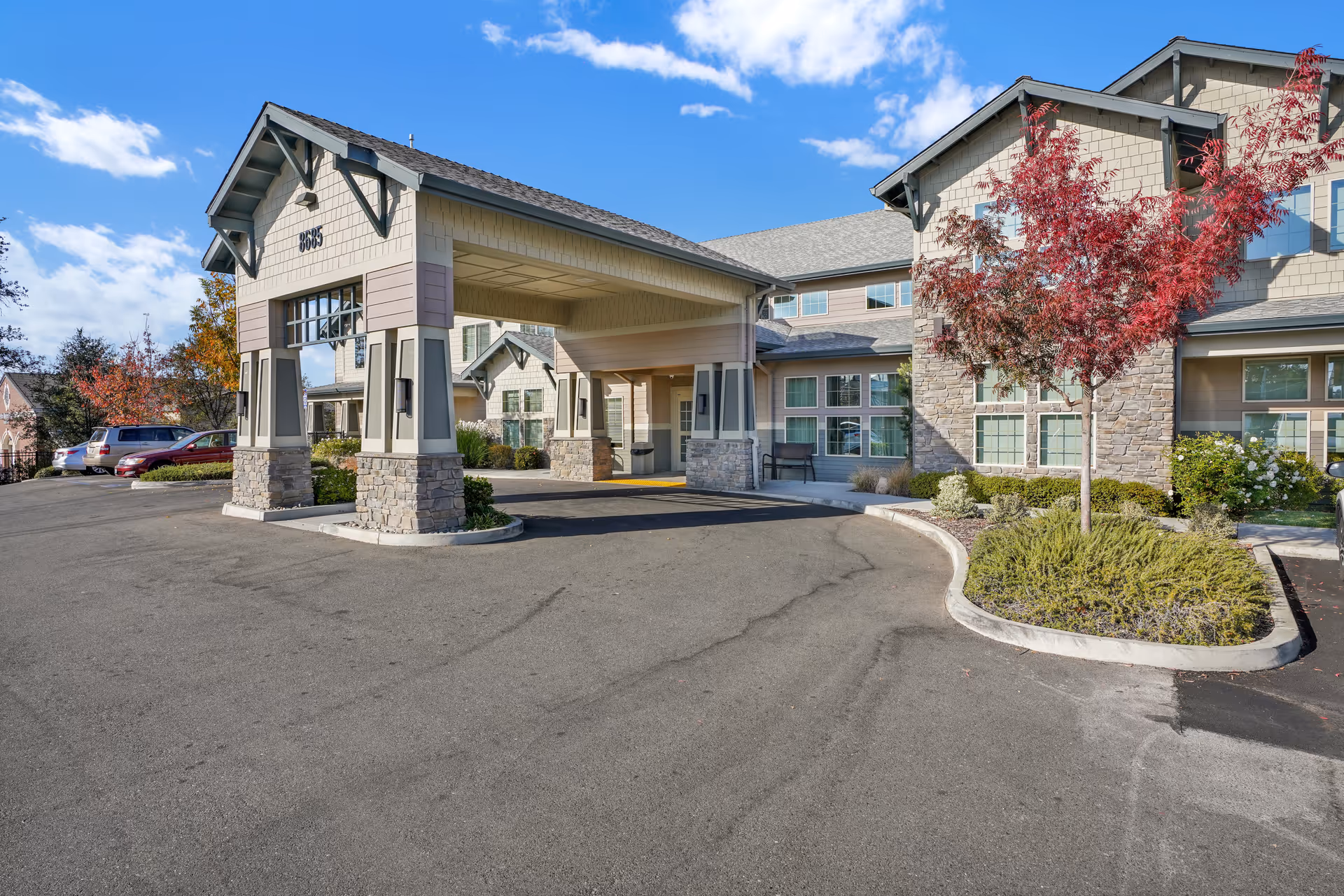 Exterior view of Almond Heights senior living facility showing the main entrance with a covered drop-off area, stone and siding facade, landscaped bushes and a small tree with red leaves, and a clear blue sky with some clouds.