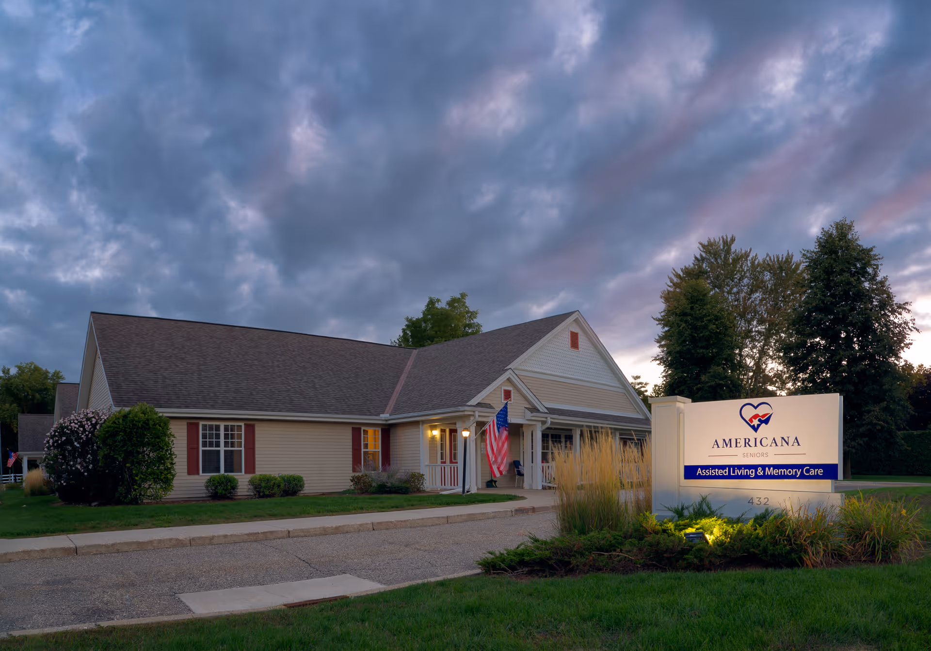 Exterior view of Americana Seniors of Davison building at dusk with a cloudy sky. The building has beige siding, a dark roof, and an American flag near the entrance. A well-lit sign in front reads 'Americana Seniors Assisted Living & Memory Care'.
