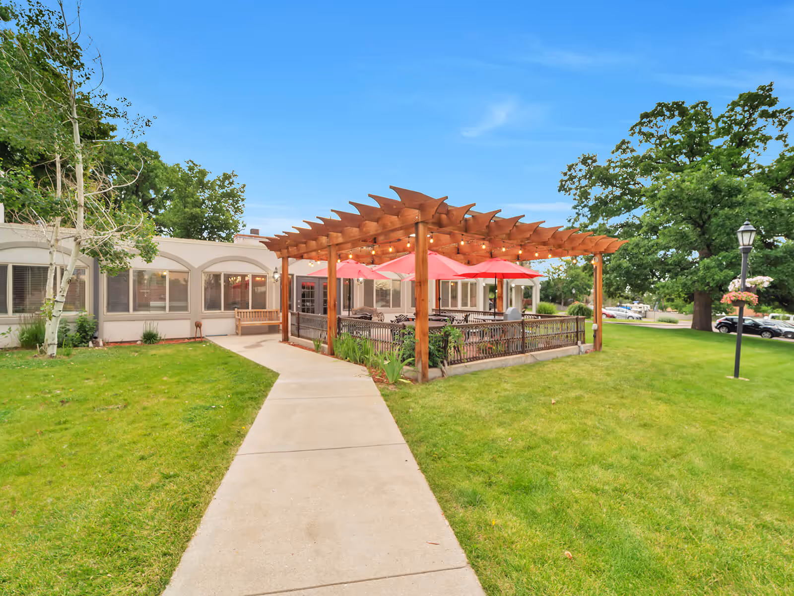Outdoor patio with a wooden pergola and red umbrellas beside a senior living building, with a concrete walkway and green lawn.