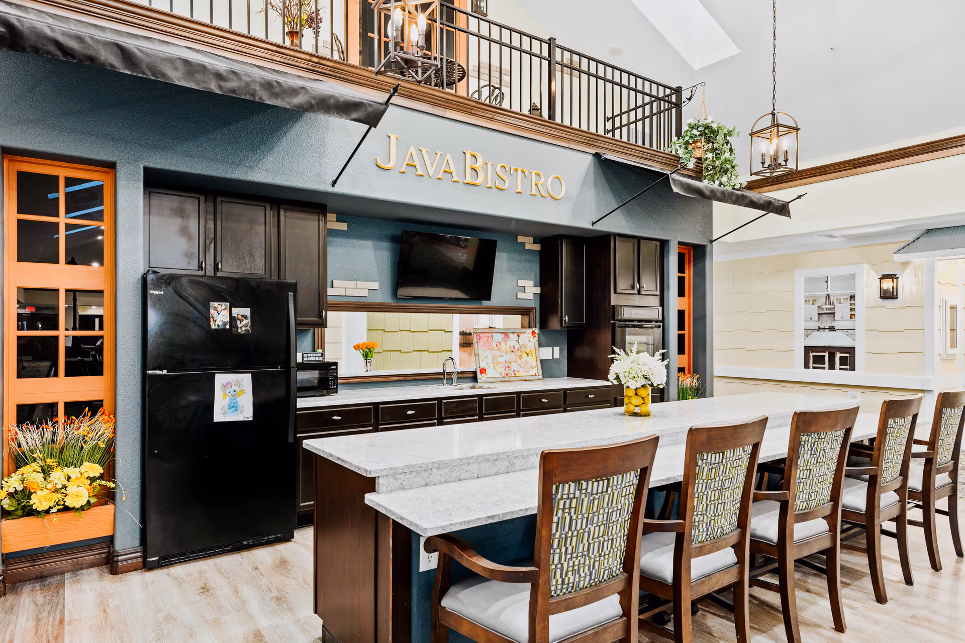Interior view of a modern kitchen area labeled 'Java Bistro' with a long island countertop surrounded by wooden chairs with patterned cushions. The kitchen features dark cabinetry, a black refrigerator with children's artwork attached, a wall-mounted TV, and decorative plants and flowers. The space has a bright and welcoming atmosphere with light wood flooring and a high ceiling.