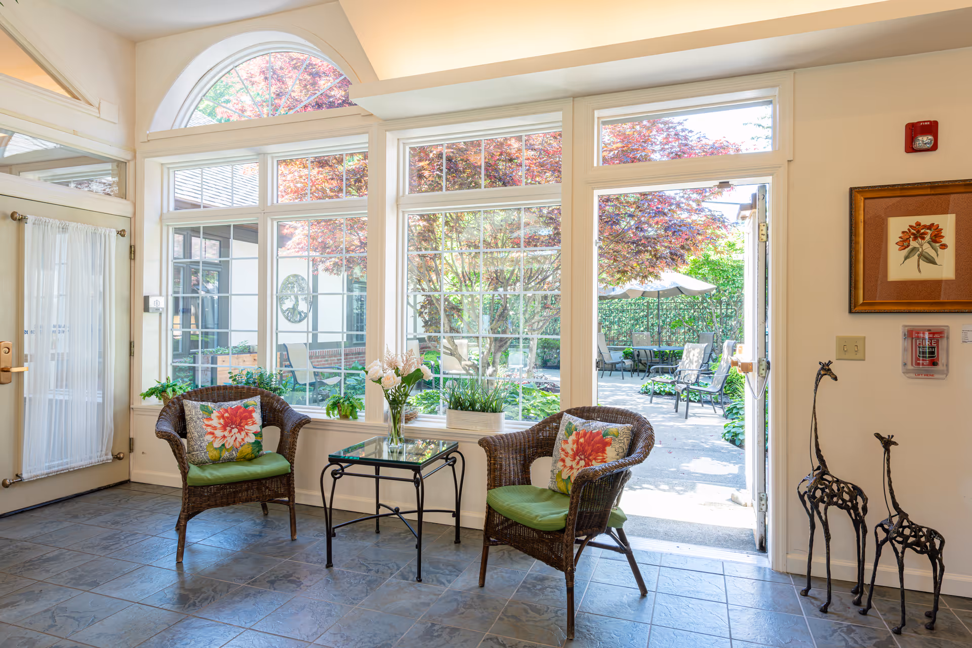 Sunlit common sitting area with two wicker chairs, a glass-top side table, and large windows and a door opening to a garden patio.