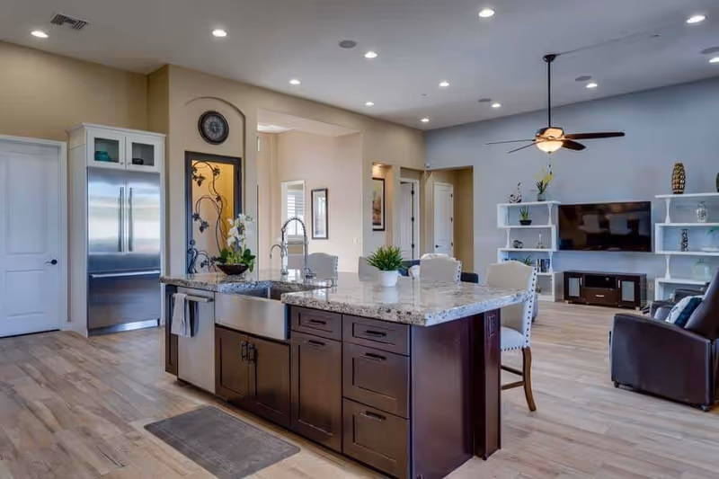 Spacious open-concept kitchen and living room area with a large granite island featuring a farmhouse sink and dark wood cabinetry. The kitchen includes a stainless steel refrigerator and light wood flooring. The living room has a ceiling fan, a mounted flat-screen TV, white shelving units with decorative items, and a dark leather armchair.