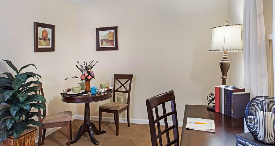 A cozy corner of a room featuring a small round wooden table set for two with plates, cups, and a floral centerpiece. Two wooden chairs with cushioned seats are placed around the table. On the right side, there is a wooden desk with a lamp, several books, and some papers with a pencil. A large green plant is positioned on the left side, and two framed pictures hang on the beige walls.