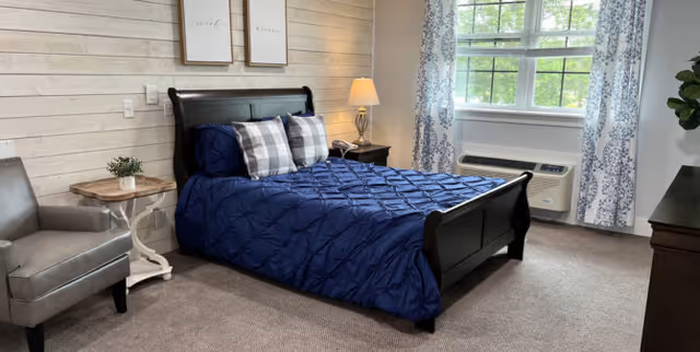 A bedroom with a dark wooden bed frame and a navy blue comforter with two plaid pillows. There is a gray armchair next to a small round wooden side table with a plant on it. A nightstand with a lamp and a telephone is beside the bed. The room has light-colored wood panel walls, two framed pictures above the bed, a window with patterned curtains, and a carpeted floor.