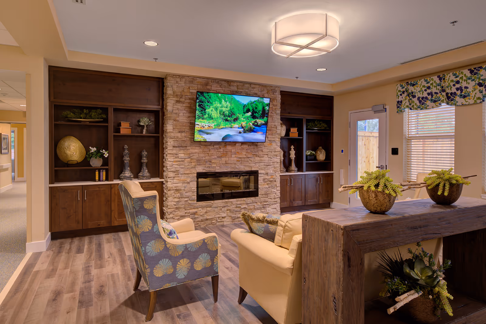 Cozy communal living room with armchairs, a stone fireplace with mounted TV, built-in wooden shelving, and decorative plants.