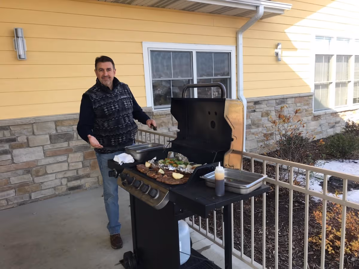 A man standing outdoors next to a gas grill with food cooking on it, including vegetables and meat. The setting is a patio area with a yellow building wall and stone accents in the background, along with some plants and a railing.