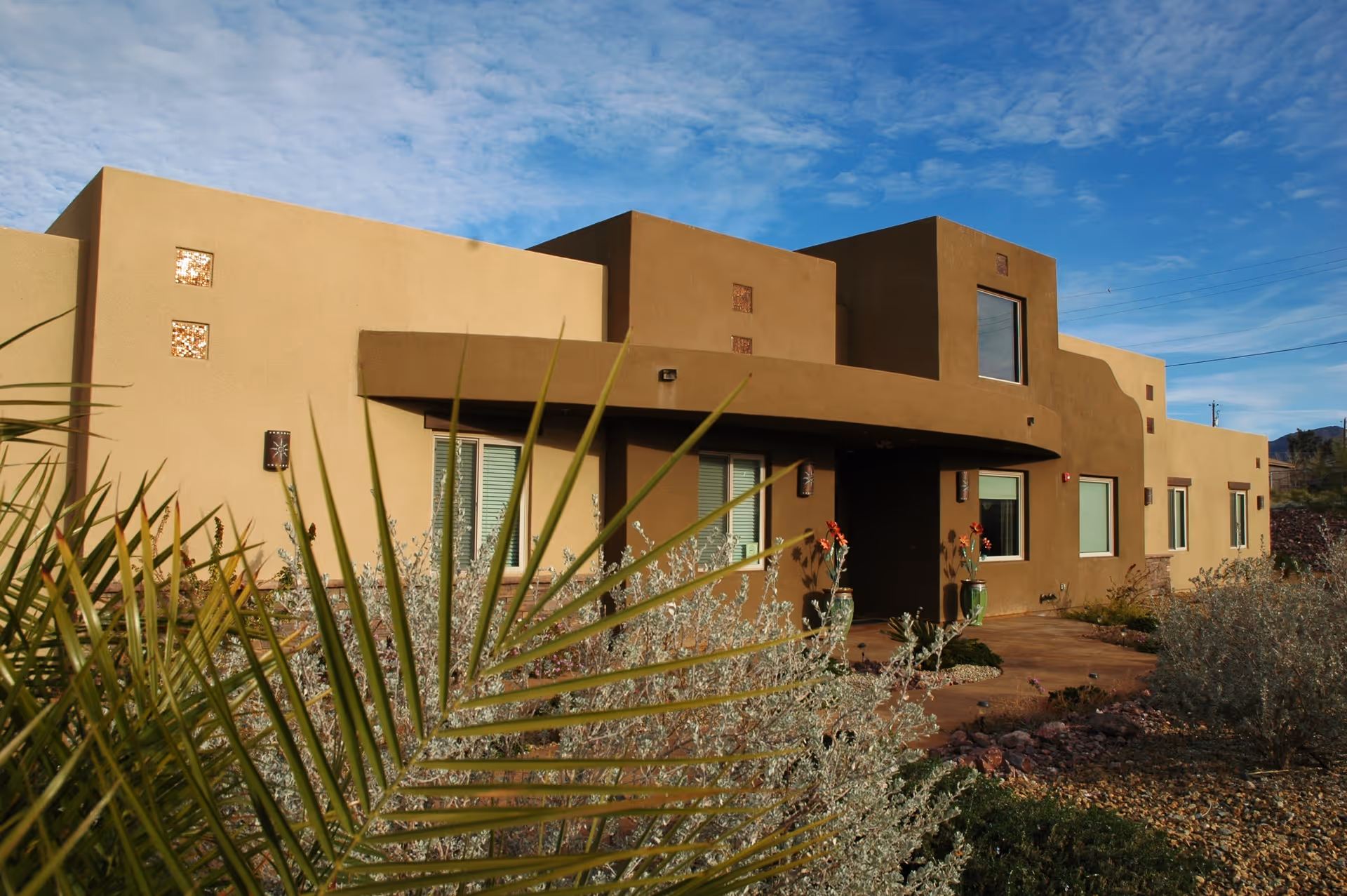 Front exterior of a modern stucco assisted living building surrounded by desert landscaping under a blue sky.