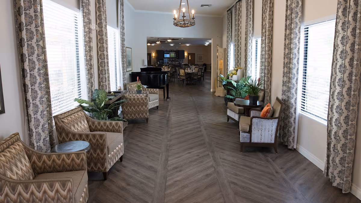 Long bright communal lounge with armchairs, potted plants, large windows, and a piano leading into a dining area.