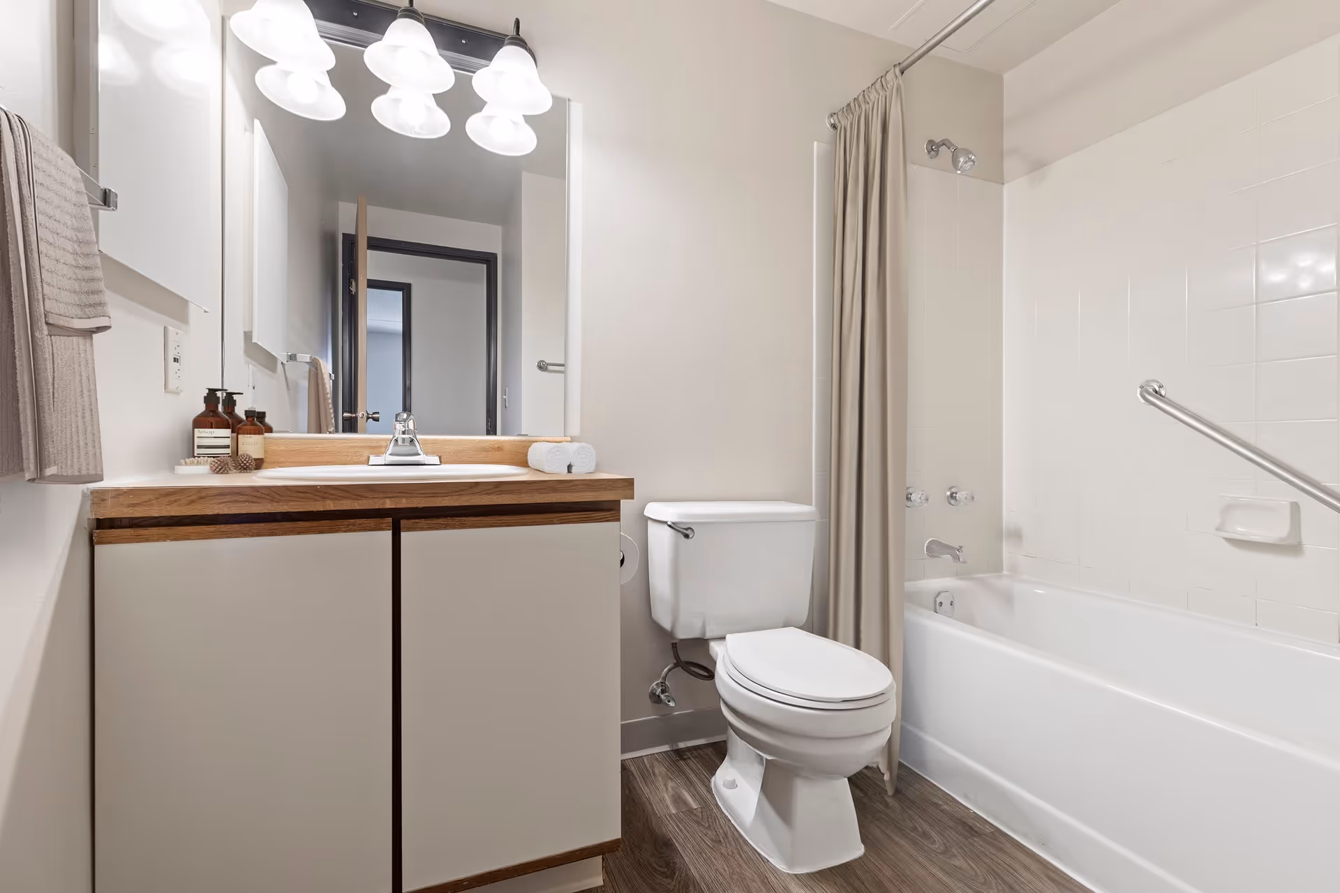A clean bathroom featuring a white toilet, a bathtub with a beige shower curtain and a grab bar, a wooden vanity with a white sink, a large mirror above the sink, and three light fixtures with frosted glass shades. There are two rolled white towels on the vanity and two brown bottles of soap or lotion next to the sink. The floor has a wood-like finish.
