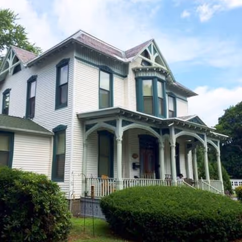 White Victorian-style senior living building with a wraparound porch and manicured hedges.