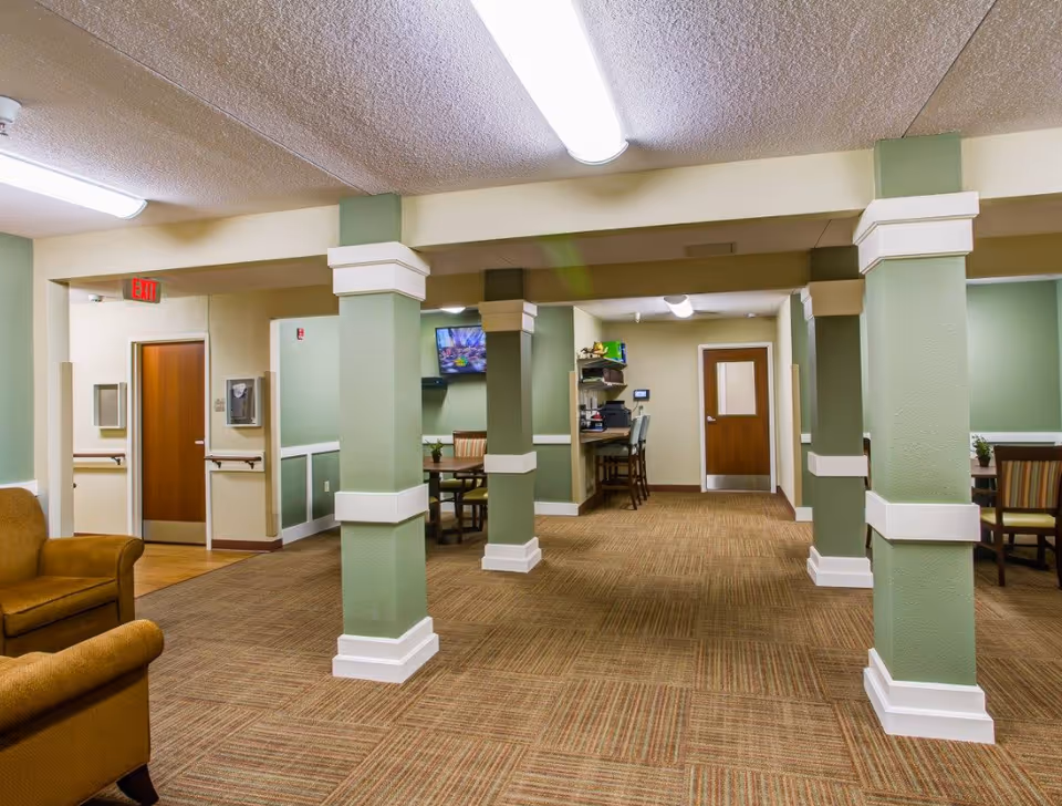 Interior view of a senior living facility common area with green and beige walls, carpeted floor, several square columns with white trim, a brown armchair on the left, a small dining table with chairs, a wall-mounted TV, and a desk with chairs and office equipment in the background.