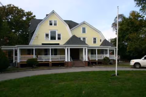 Yellow two-story building with wraparound porch and a driveway in front.