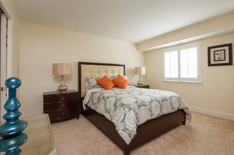 A neatly made bed with a patterned gray and white comforter and two orange accent pillows in a bright bedroom. The bed has a padded beige headboard and is flanked by two dark wooden nightstands, each with a lamp. There is a window with white shutters on the right wall and a framed picture hanging beside it. A blue decorative glass object is visible on a piece of furniture in the foreground on the left.